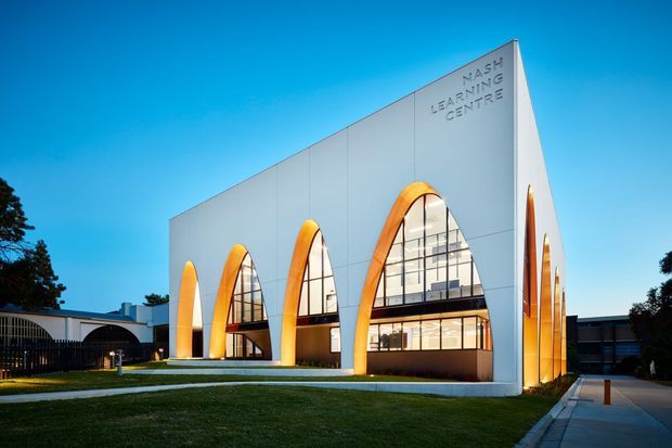 Exterior architectural photograph of a contemporary learning centre, featuring a crisp white geometric facade with a series of tall, repeated arched openings edged in warm orange. The arches contain expansive glazed curtain walls revealing interior circulation and lighting. The building sits beside a landscaped lawn and paved pedestrian approach under a clear blue sky, with signage reading “NASH LEARNING CENTRE.”