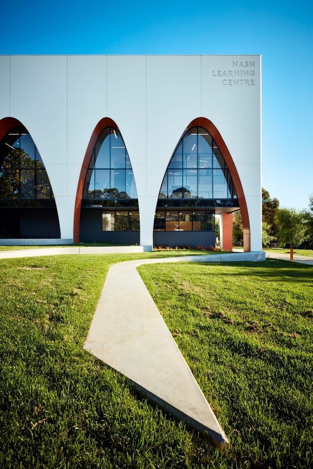 Exterior view of the Nash Learning Centre, a contemporary institutional building with a white panelled facade, large arched floor-to-ceiling glazed openings framed by reddish-brown curved structural elements, and visible interior levels. A concrete pedestrian path crosses a broad lawn in the foreground under a clear blue sky.