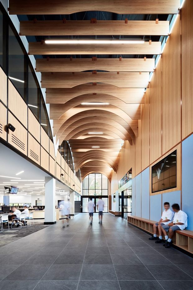 A large contemporary institutional interior resembling a school or civic building, viewed along a generous central circulation hall. The space features a dramatic repeating curved timber ceiling with exposed structural fixing details, integrated linear lighting, timber wall panelling, dark grey tiled flooring, extensive black-framed glazing, built-in timber bench seating, and an open collaborative workspace visible to the left. The corridor terminates in a tall arched glazed opening, creating a bright, warm, highly articulated interior.