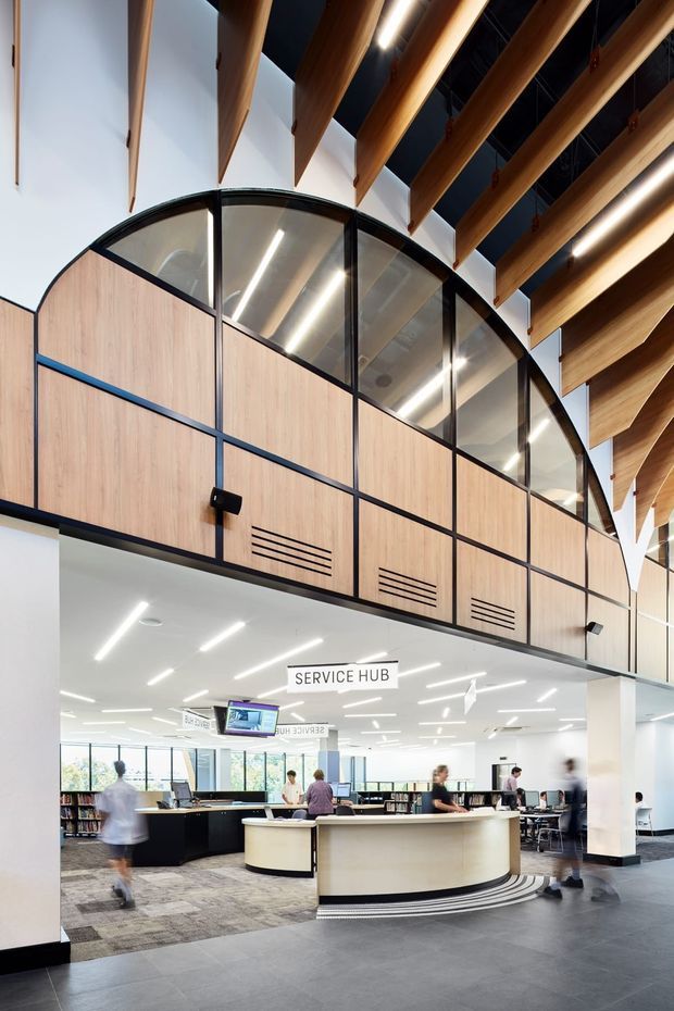 A bright contemporary service hub in a large institutional interior, featuring a curved reception counter, open-plan work and library areas, pale timber and black-framed partitioning, clerestory glazing, and dramatic exposed timber roof beams with linear lighting. The space has a polished, functional public-building character with high ceilings and strong structural detailing.