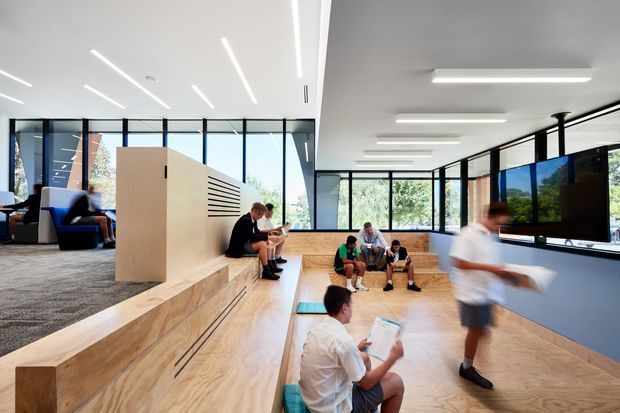 Bright contemporary communal interior with an open-plan stepped timber seating area, large black-framed glazed walls, integrated bench seating, and linear recessed lighting. Several people occupy the tiered space, which appears suited to collaborative work, learning, or informal gathering. A carpeted lounge zone with armchairs is visible at left, while the main room has warm timber flooring and pale walls.