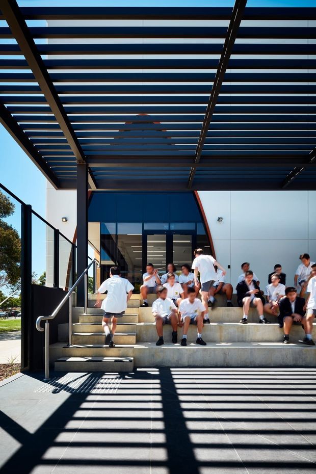 Exterior architectural view of a contemporary institutional or commercial building entrance. Broad concrete steps and a dark paved forecourt sit beneath a large slatted pergola or canopy that casts strong linear shadows. The facade combines white panel cladding, dark framing, extensive glazing, and a red-edged angular geometric feature. People are seated on the steps, emphasizing the building's public entrance and substantial scale.
