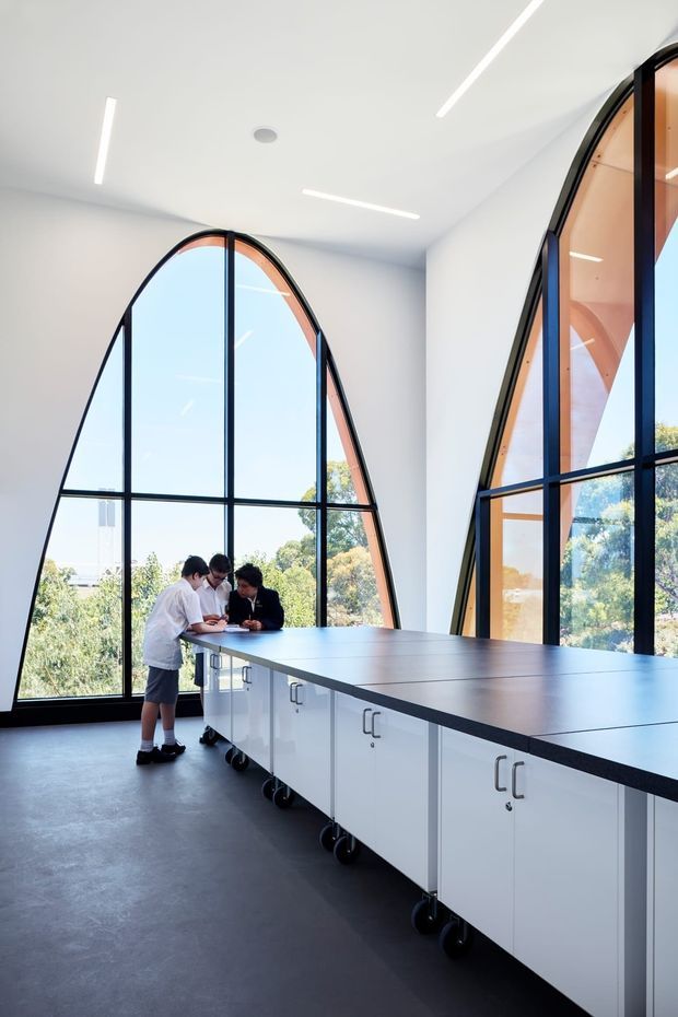Bright contemporary interior featuring a long white built-in island or workbench with mobile storage cabinets and a dark benchtop. The room has dramatic arched floor-to-ceiling windows with black metal frames, white walls and ceiling, linear recessed lighting, and a dark polished floor. Several people are gathered at the workstation, suggesting a collaborative studio, classroom, or commercial workspace.