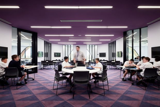 A large open-plan commercial lounge or breakout area with multiple round tables, black chairs, and people working or dining. The space features extensive internal glass partitions with black frames, white walls, a striking purple ceiling, geometric purple and blue patterned carpet, wall-mounted screens, and evenly spaced linear LED lighting. The symmetrical layout and communal seating suggest a contemporary workplace, education, or hospitality setting.