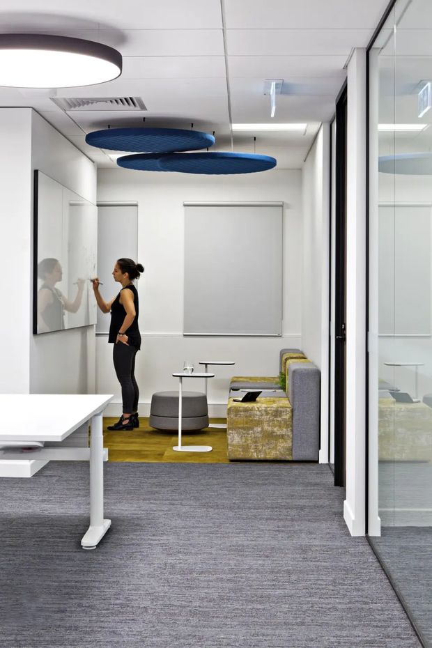 A bright, modern commercial office interior with an open workspace in the foreground and a compact meeting or breakout area beyond. The room has white walls and ceiling panels, grey carpet flooring, glass-partitioned rooms on the right, a white worktable, and a lounge setting with grey upholstered seating, small white tables, and a muted yellow-green rug. Blue acoustic ceiling discs and simple contemporary lighting provide visual accents. A person is writing on a wall-mounted whiteboard, with her reflection visible in the adjacent glass.