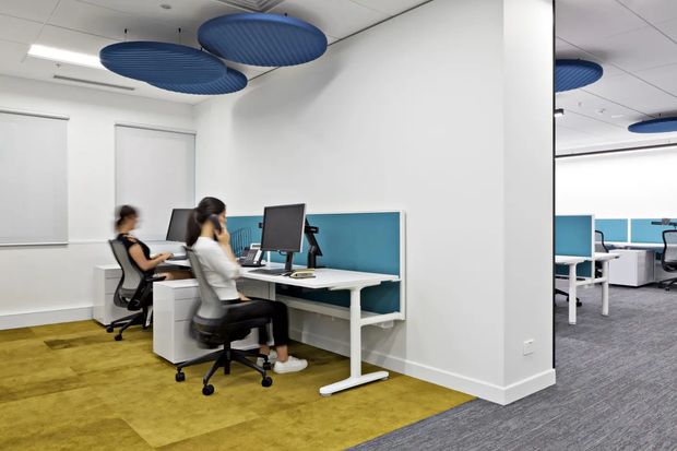 A bright contemporary open-plan office with paired workstations, white desks, blue privacy screens, office chairs, computer monitors, and acoustic blue circular ceiling panels. The space has white walls and a mix of mustard-toned carpet tiles and grey carpet, with a partial office area visible in the large wall mirror or glazed reflection.