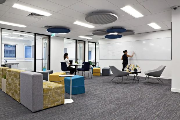 Contemporary open-plan commercial collaboration and breakout area with modular upholstered seating, small tables, glass-partitioned meeting rooms, a large whiteboard wall, carpeted flooring, and suspended acoustic ceiling panels. Bright linear LED lighting and a neutral grey palette create a functional, modern office environment.