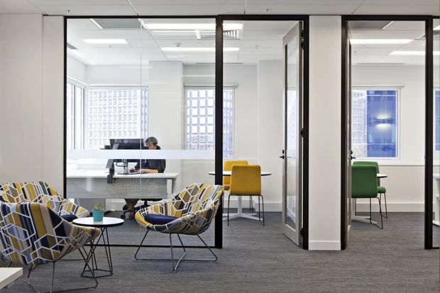 A bright contemporary commercial office interior viewed from a lounge or reception area. The foreground contains patterned upholstered lounge chairs and small round tables, while glazed black-framed partitions enclose open workspaces with desks and colourful task chairs. Grey carpet, white walls, suspended acoustic ceiling tiles, integrated fluorescent lighting, and glass-block windows create a clean, functional workplace environment.