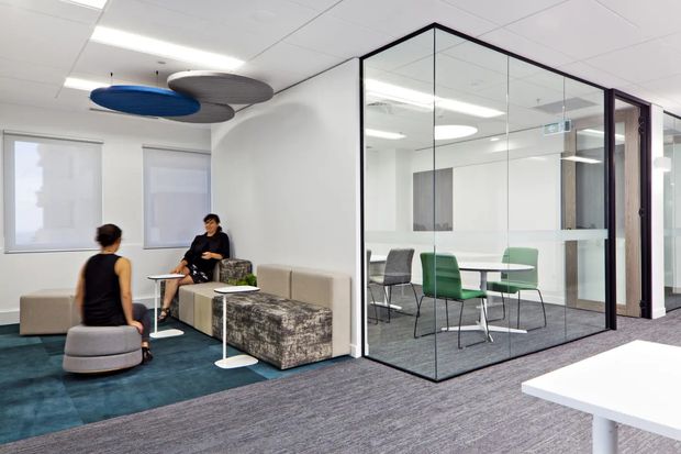 A bright contemporary commercial office interior with an informal lounge and meeting area. The open workspace includes upholstered modular seating on a dark teal carpet, a glass-walled meeting room with a white table and green and grey chairs, white acoustic ceiling tiles, and colourful suspended acoustic ceiling panels. Black-framed glazing and a dark timber door add definition to the meeting enclosure.