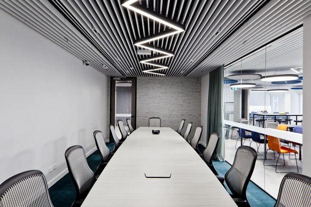 Modern commercial meeting room with a long white conference table and ergonomic mesh chairs. The space features a linear slatted ceiling with a striking zigzag LED light fixture, a textured grey feature wall, dark teal carpet, glazed partitions, and a visible open office area beyond.
