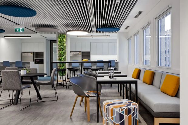 A bright contemporary communal office or hospitality-style breakout space with multiple dining and work tables, upholstered chairs, a long built-in bench seat with mustard cushions, a compact kitchenette, and a blue-accented linear acoustic ceiling. Large windows provide abundant daylight, while indoor planting and colourful geometric furniture add visual interest.
