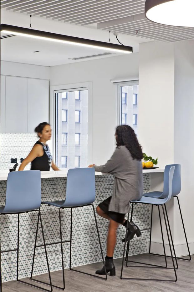 Bright contemporary commercial-style kitchen or breakout area with a long island/breakfast bar, pale blue bar stools, white cabinetry, and a geometric black-and-white tiled island face. The space has large windows, a white slatted ceiling, linear pendant lighting, and grey timber-look flooring. Two people are seated and standing at the counter.