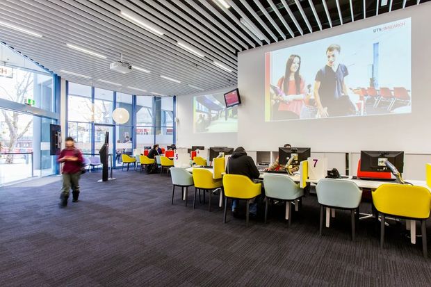 Bright modern commercial learning or study space with rows of individual workstations, computer monitors, colorful yellow and grey chairs, large projection screens, extensive glazing, and a linear slatted ceiling. The open-plan interior has a contemporary institutional or educational character.