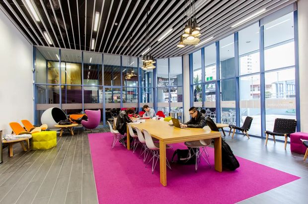 A bright, contemporary open-plan commercial workspace or breakout area with a long communal timber table, multiple colourful chairs, lounge seating, and a vivid magenta rug. The space features floor-to-ceiling blue-framed glazing, an exposed linear slatted ceiling with integrated lighting, and polished dark flooring. Urban surroundings are visible through the windows.