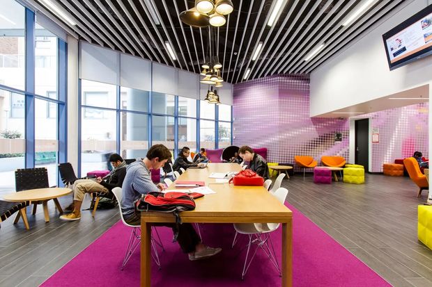 A bright contemporary commercial lounge or coworking space with large blue-framed windows, a long timber communal table, colourful seating, magenta carpet runners, and a striking pink mosaic-tile feature wall. The ceiling features dense black-and-white linear slats with suspended decorative pendant lights. People are working at the shared table, and a wall-mounted screen is visible.