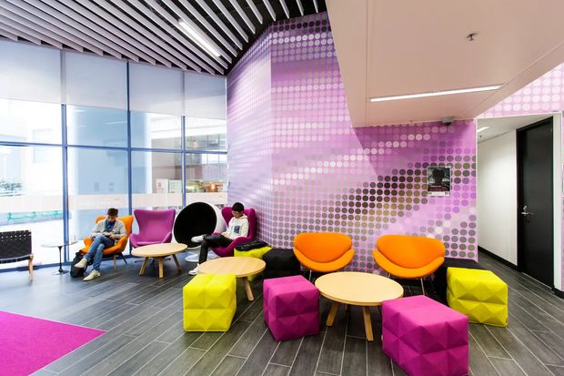 A bright contemporary commercial lounge or breakout area with floor-to-ceiling glazing, dark tiled flooring, colourful lounge chairs and faceted ottomans, round timber tables, and a vivid purple circular-pattern feature wall. A linear white ceiling light and a slatted ceiling zone add architectural interest.