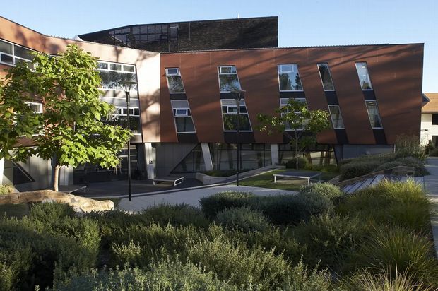 Exterior view of a contemporary multi-storey institutional or commercial building, featuring a dark brick upper volume, rust-brown metal-clad facade panels, irregularly angled vertical windows, and a landscaped courtyard with native-style planting and paved walkways.