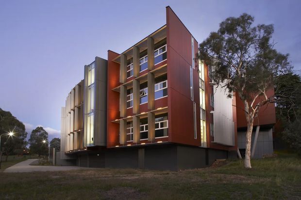 Exterior view of a contemporary multi-storey building with a strongly articulated facade. The central elevation combines dark grey vertical fins, warm red-brown metal or panel cladding, and rows of horizontal windows, with lighter silver-grey side volumes. The building sits in a landscaped suburban or institutional setting and is photographed at dusk.