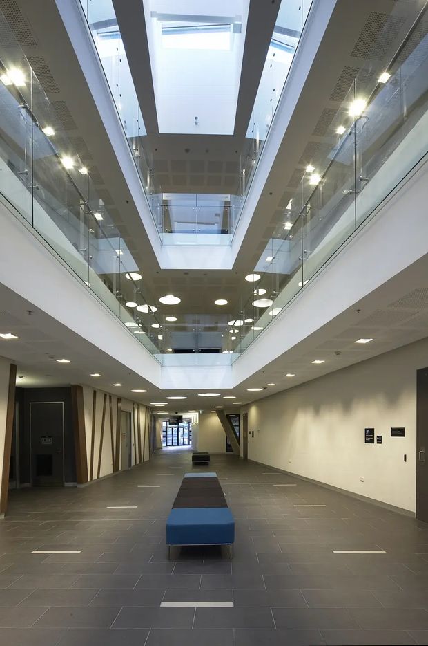 A bright contemporary institutional or commercial interior hallway with a long central corridor, dark tiled flooring, white walls, recessed lighting, glass balustrades, and a dramatic multi-level atrium. Large overhead skylights illuminate the open void, while blue and charcoal upholstered benches form a central seating arrangement. The space has clean, functional detailing and a spacious, airy character.