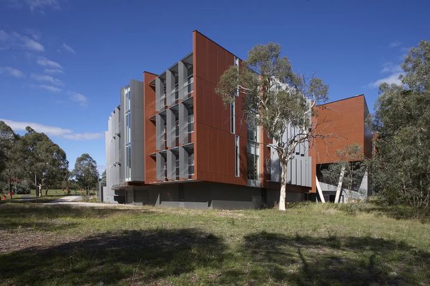 Exterior view of a contemporary multi-storey building set within a landscaped, tree-lined site. The facade combines large rust-red metal-clad wall planes with contrasting light grey vertical elements, recessed balconies or screened openings, extensive glazing, and a dark raised base. The composition is angular and sculptural, with mature eucalyptus-like trees and open grass surrounding the building under a clear blue sky.