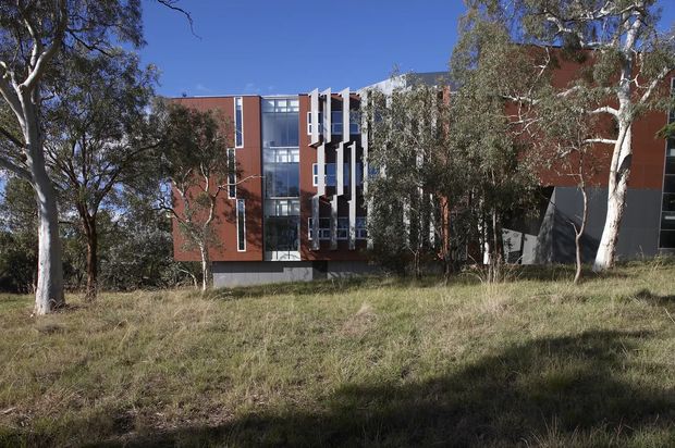 Exterior view of a contemporary multi-storey building partially screened by native trees and dry grass. The facade combines red-orange wall panels, dark grey sections, extensive vertical glazing, and projecting white structural or window-frame elements against a clear blue sky.