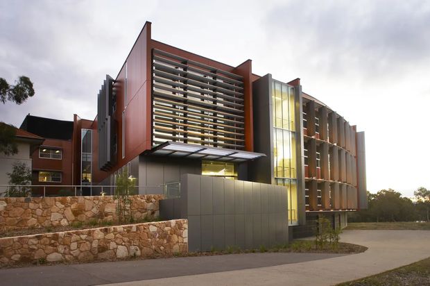 Exterior view of a contemporary multi-storey building with strong geometric massing, reddish-brown and grey cladding, extensive horizontal sun-shading louvres, and a tall glazed vertical atrium or stairwell. The building sits behind landscaped stone retaining walls and a paved forecourt, with a smaller older building visible to the side.