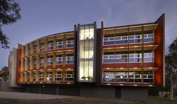 Exterior dusk view of a contemporary multi-storey commercial or institutional building. The facade features extensive horizontal window bands, vertical structural fins, warm recessed lighting, dark lower-level cladding, and a prominent glazed central circulation or atrium tower.