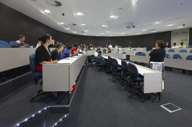 A large tiered lecture theatre or seminar room with curved rows of fixed desks, blue upholstered seating, wheeled office chairs, and numerous attendees. The room has dark acoustic wall panels, a light suspended ceiling with recessed lighting and ventilation grilles, and dark carpet flooring. A projection system and wall-mounted equipment are visible at the front or side of the room.