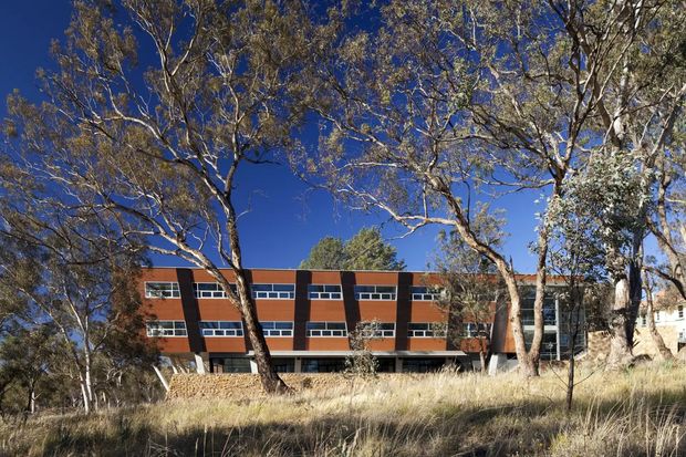 Exterior view of a low-rise modern building set among mature eucalyptus trees and dry native grassland. The facade combines reddish-brown brick or masonry panels, dark vertical structural elements, and long horizontal bands of glazing, with a glazed entrance or stairwell visible at the right. The clear blue sky and bushland setting suggest an Australian suburban or semi-rural campus environment.