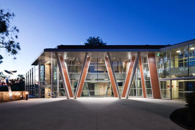A modern multi-storey institutional or commercial building photographed at dusk. The broad glazed facade features prominent angled structural columns forming repeated V-shaped supports, a dark shallow roofline, exposed interior circulation spaces, and a large paved forecourt. The architecture has a crisp contemporary civic or educational character.