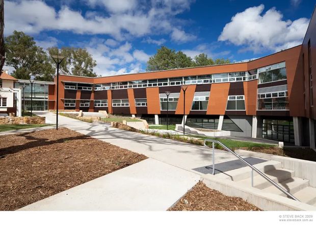 Exterior architectural photograph of a contemporary multi-storey institutional or commercial building arranged around a landscaped courtyard. The facade combines dark red-brown brick or masonry, extensive horizontal glazing, and projecting orange-red metal or panel elements. A broad concrete pedestrian path, stairs, stainless-steel handrails, low retaining walls, and young landscaping lead toward the building. Mature trees, blue sky, and a small portion of an adjacent older building are visible in the background.