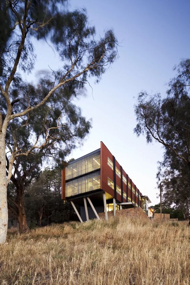 Exterior architectural photograph of a contemporary multi-storey building set among mature trees and dry grassland. The building features a long cantilevered rectangular form, dark-framed floor-to-ceiling glazing with vertical screening, red-brown solid wall panels, and angled structural supports beneath the elevated upper levels. The scene is photographed in soft daylight with a natural, semi-rural setting.