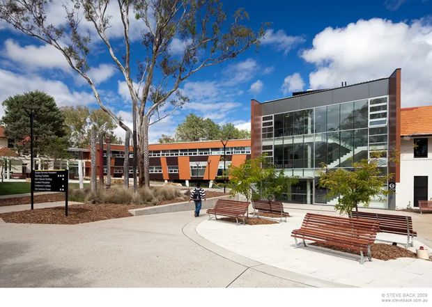 Exterior view of a contemporary multi-storey institutional or commercial building surrounding a landscaped pedestrian courtyard. The complex combines red brick and dark grey cladding with extensive floor-to-ceiling glazing, visible internal stairs, long horizontal window bands, and a red tiled roof on an adjoining section. Paved paths, timber benches, mature trees, and directional signage occupy the foreground under a bright blue sky.