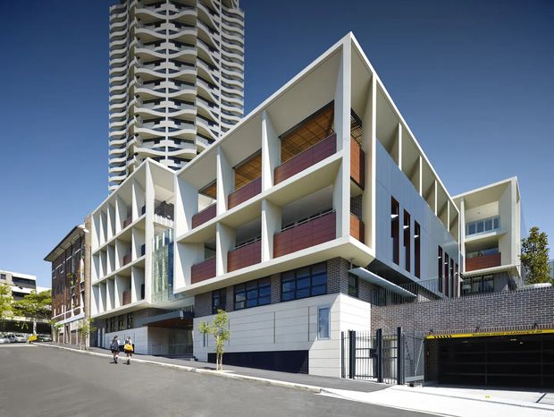 Street-level exterior view of a contemporary multi-storey urban development, combining a sculptural white low- to mid-rise building with recessed balconies, projecting fins, dark brickwork, and a taller residential tower behind it. The facade includes red-toned balcony screens, extensive glazing, a gated basement garage entrance, and geometric facade articulation under a clear blue sky.