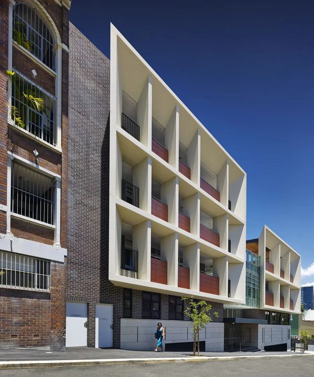 Street-level exterior view of a contemporary multi-storey residential building integrated beside an older brick structure. The new facade features a strong geometric grid of projecting balconies or recessed terraces, cream-white concrete framing, dark metal balustrades, and red-toned privacy panels. A glazed vertical circulation or lobby section connects the main wing to a smaller stepped wing. The image is taken in an urban setting under a clear blue sky.
