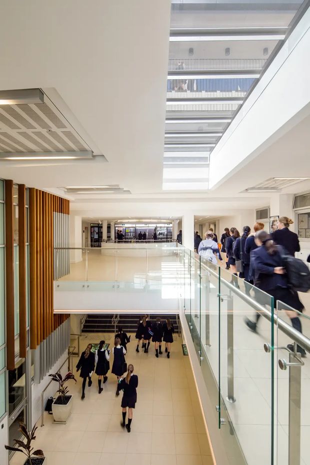 Bright multi-level institutional interior, likely a school or educational building, viewed from an upper-level corridor overlooking a central atrium. The space features glass balustrades, a large overhead skylight, pale tiled flooring, white walls and ceilings, linear ceiling lighting, and a prominent vertical timber slat screen. Students and staff move along the corridors and below.