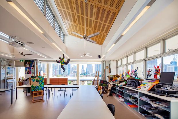 A bright, spacious art studio or creative classroom with long white worktables, stools, open shelving filled with art supplies, easels, colourful artwork, and large windows overlooking a city skyline. The room has exposed acoustic timber ceiling panels, ceiling fans, linear lighting, and extensive glazing, creating an airy collaborative workspace.