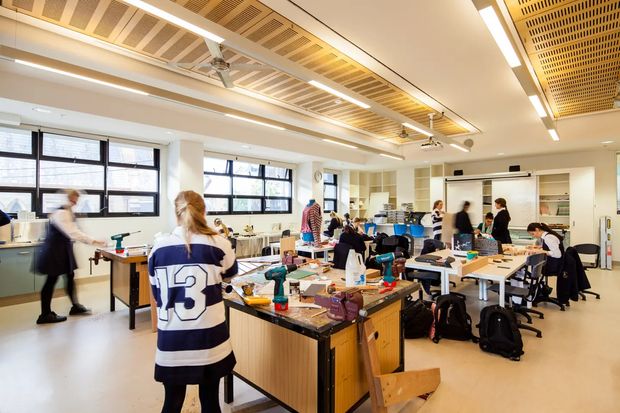 Bright, spacious creative workshop or design studio with multiple shared worktables, stools, tools, craft materials, computers, and people working. The room has large black-framed windows, white walls, pale flooring, exposed services, ceiling fans, linear lighting, and prominent timber slatted acoustic ceiling panels. It appears to be an educational or commercial makerspace rather than a residential room.