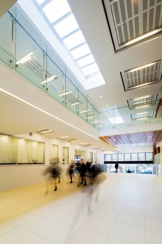 A bright, spacious multi-level commercial interior or institutional atrium with a large tiled floor, reception or service counter, glass balustrades on an upper mezzanine, extensive skylights, and exposed ceiling services. The space has white walls and floors, strong linear lighting, visible HVAC grilles, and blurred people moving through the lobby. A timber-lined ceiling feature adds warmth near the rear of the atrium.