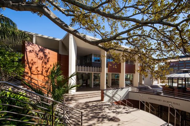 Exterior view of a contemporary multi-level institutional or commercial building framed by mature trees and tropical planting. The facade combines warm terracotta-coloured wall panels, pale rendered structural columns, shaded glazing, vertical screening, broad stairs, ramps and metal handrails. A sign identifies the building as a hall, with a covered outdoor walkway or courtyard visible to the right.