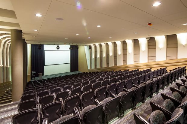 A large auditorium or lecture theatre viewed from the rear, with many rows of dark upholstered seats arranged in a steeply raked layout facing a projection screen and curtained stage. The room has a low suspended ceiling with recessed lighting, warm timber-toned balcony or wall trim, patterned carpet, and distinctive curved illuminated wall recesses along the side walls.