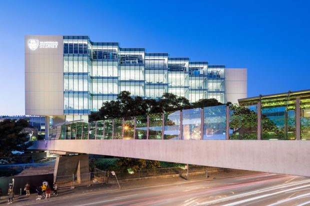 Exterior dusk view of a contemporary multi-storey university or institutional building with extensive glass curtain-wall glazing, pale solid wall sections, and a stepped building profile. A landscaped elevated pedestrian structure with planted trellises crosses the foreground above a busy roadway with light trails.