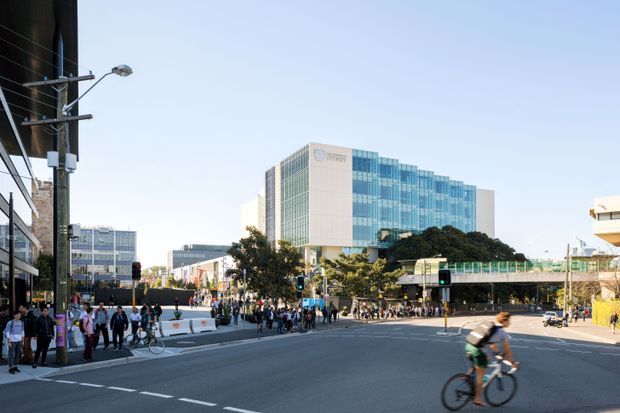 Wide urban streetscape showing a modern multi-storey commercial building with extensive blue-green glazed curtain walling, a busy intersection, pedestrians, cyclists, street lighting, and surrounding city buildings. The image is an exterior architectural and urban context view in clear daylight.