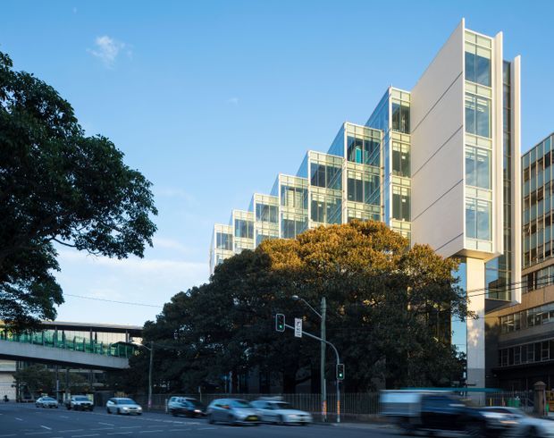 Street-level view of a contemporary multi-storey commercial or institutional building with a stepped sequence of glazed volumes, tall narrow window bays, and a prominent pale solid facade. The building fronts a busy urban road with traffic lights, mature street trees, and a pedestrian overpass in the foreground.