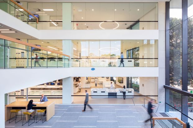 Bright multi-level commercial interior with open-plan workspaces, glass balustrades, extensive floor-to-ceiling glazing, white structural bands, and a central atrium. The ground level includes collaborative tables and seating, while upper levels show offices and circulation areas. Contemporary circular pendant lighting and pale finishes create an airy, professional workplace atmosphere.