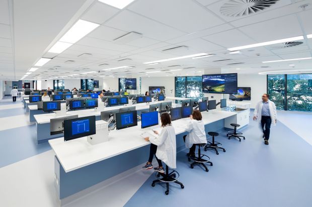 Bright, expansive modern commercial operations or control-room interior with rows of white workstation desks, multiple computer monitors, ergonomic office chairs, large wall-mounted display screens, extensive glazing, and a suspended acoustic ceiling with integrated fluorescent lighting and air-conditioning grilles. The space has a clean white and pale blue fit-out and appears designed for collaborative technical or monitoring work.
