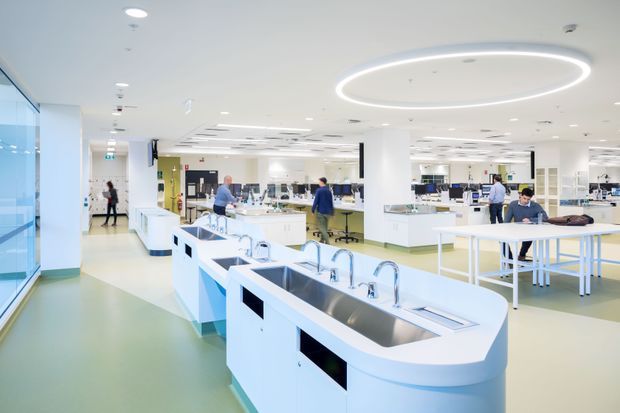 Bright, spacious contemporary commercial interior resembling a laboratory, educational science facility, or technical workspace. The open-plan room has white built-in workbenches with integrated sinks and curved edges, numerous taps, laboratory equipment, stools, desks, computers, and people working throughout. A large circular LED pendant feature is set into the white ceiling, with extensive recessed lighting, pale green flooring, white columns, and glazed walls.