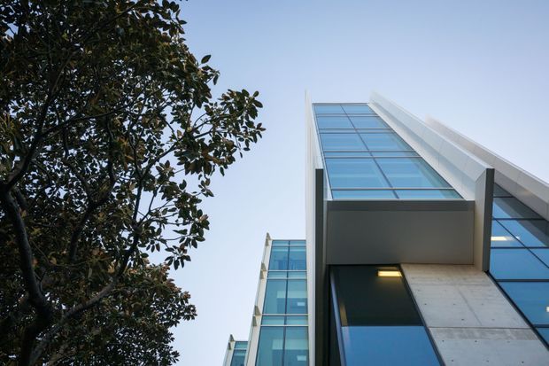 Low-angle exterior view of a contemporary multi-storey building, featuring extensive blue-tinted floor-to-ceiling glazing, crisp white projecting roof or facade elements, and exposed concrete wall panels. A mature tree frames the left side against a clear sky.