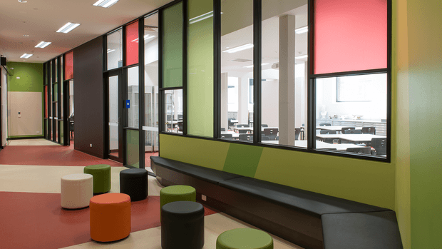A contemporary commercial interior showing a colourful school or office corridor with glazed meeting or classroom spaces on one side. The corridor includes built-in dark bench seating, small cylindrical stools in orange, green, white and black, bold green and red wall panels, black-framed internal glazing, and linear fluorescent ceiling lights. Bright workspaces are visible through the glass.