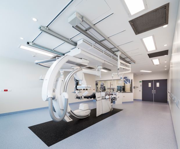 Bright, clean clinical imaging and interventional procedure room dominated by a large ceiling-mounted C-arm system, operating table, medical monitors, equipment booms, and adjustable examination lights. The room has pale walls, a light speckled resilient floor, white acoustic ceiling panels with recessed rectangular lighting, dark double doors, and a glazed observation or control area at the rear.